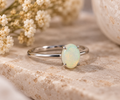 A sterling silver ring with a round, bezel-set iridescent opal gemstone, resting on a textured beige stone slab with dried white flowers in the background.