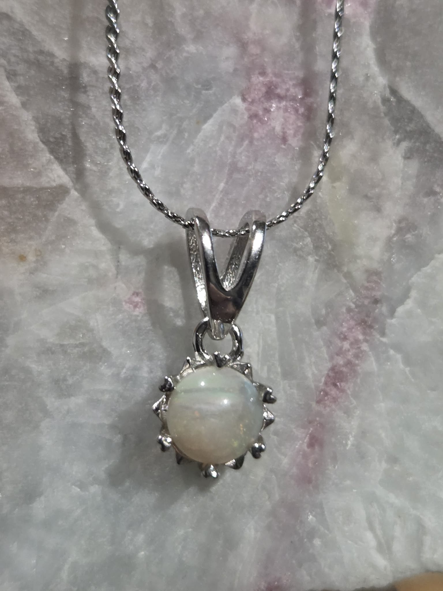 A silver pendant necklace featuring a round, iridescent white opal gemstone surrounded by a starburst-like silver setting, hanging from a twisted silver chain against a textured white crystal background.