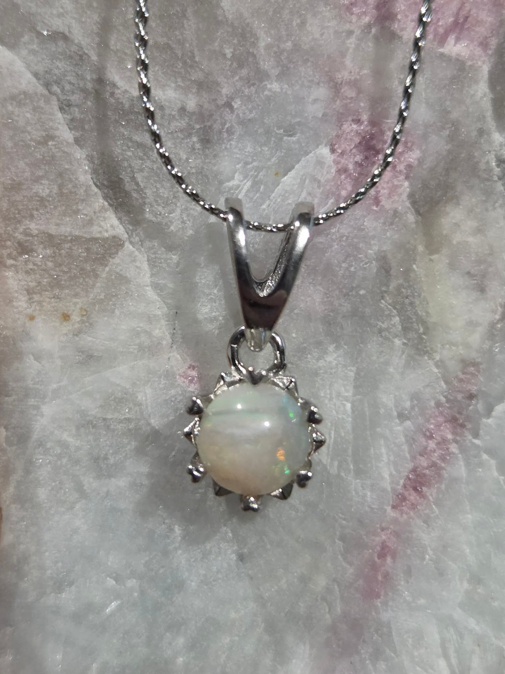 A silver pendant necklace featuring a round white opal gemstone with iridescent flashes, set in a prong-style silver frame on a matching silver chain, displayed on a textured white crystal surface.