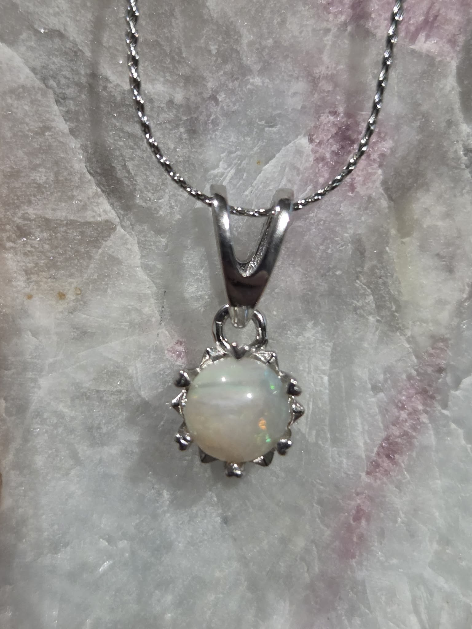 A silver pendant necklace featuring a round white opal gemstone with iridescent flashes, set in a prong-style silver frame on a matching silver chain, displayed on a textured white crystal surface.
